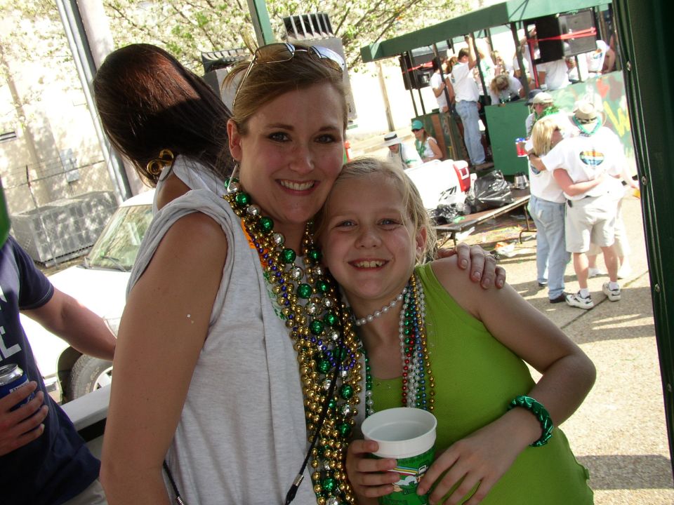 Two smiling women wearing Mardi Gras beads, with a crowd and a float in the background