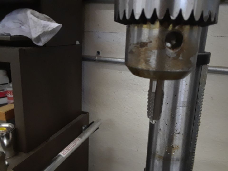 Drill press chuck holding a tap, with a rag on a shelf in the background