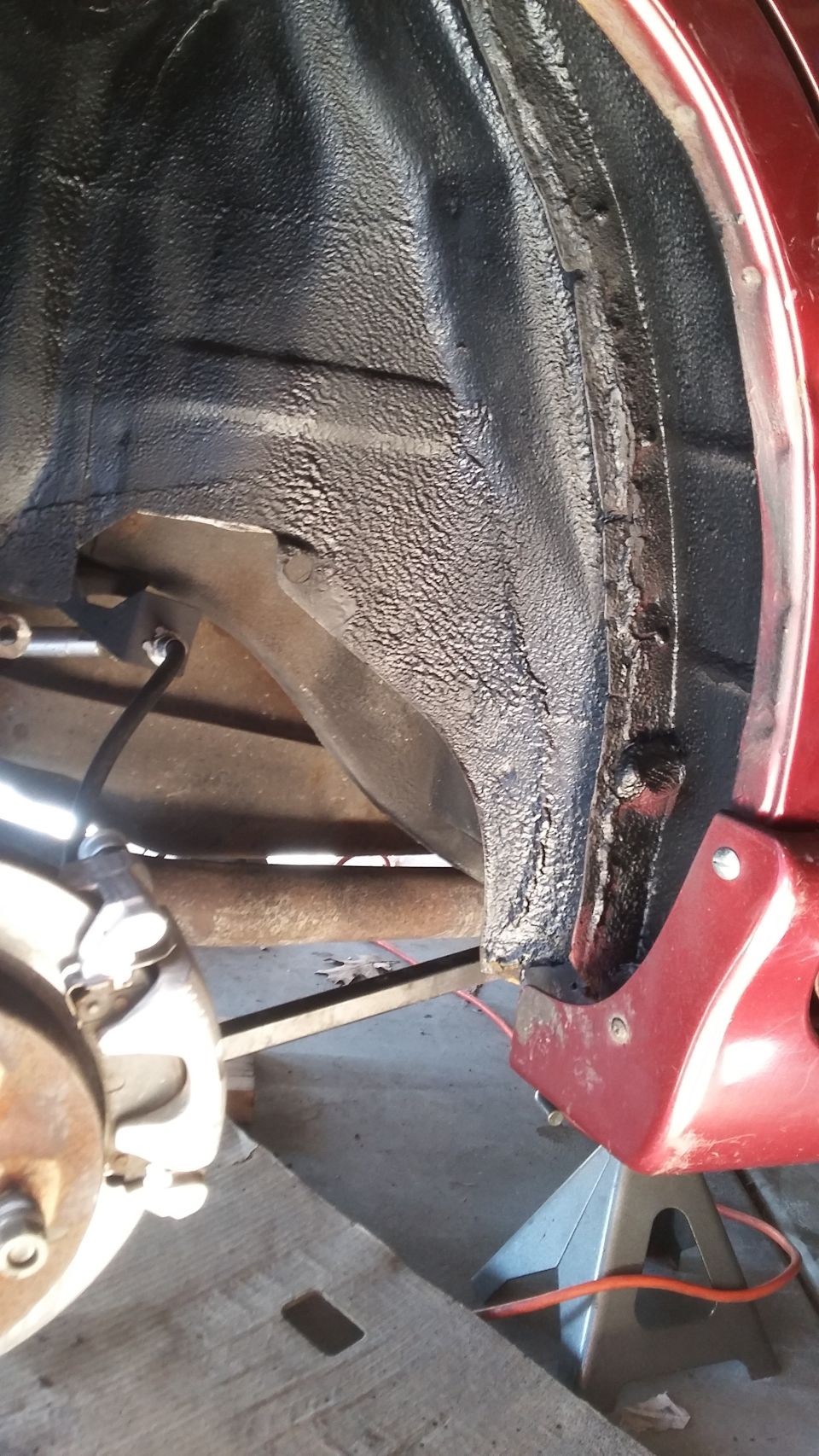 Red car's rear wheel well interior with black undercoating, jack stand, and brake components visible