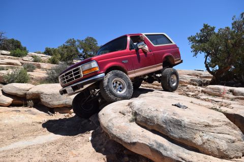 1995 Ford Bronco - A red Ford Bronco navigates a rocky off-road trail under a clear blue sky.