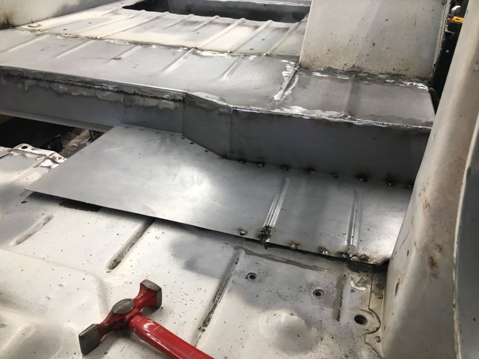Vehicle floor pan being repaired with new metal panels welded in place. A red hammer is visible on the floor