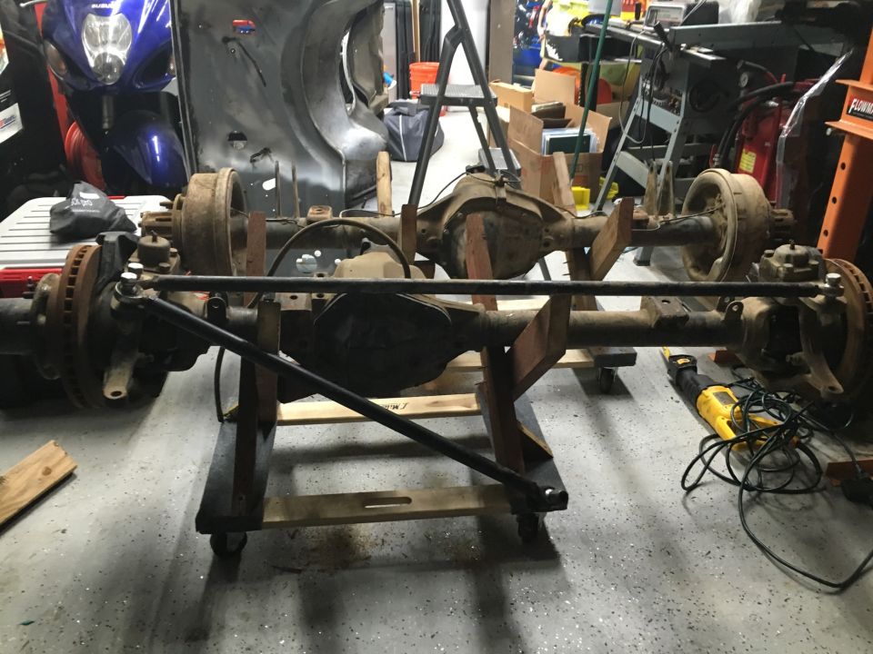 Two vehicle axles on a wooden stand, with a yellow saw on the floor