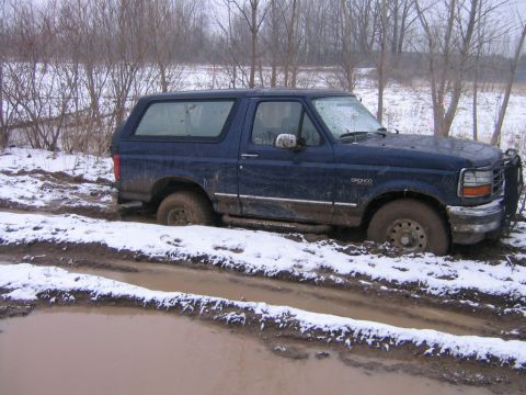 1994 Ford Bronco - A blue 1994 Ford Bronco is stuck in deep mud and snow, with trees in the background.