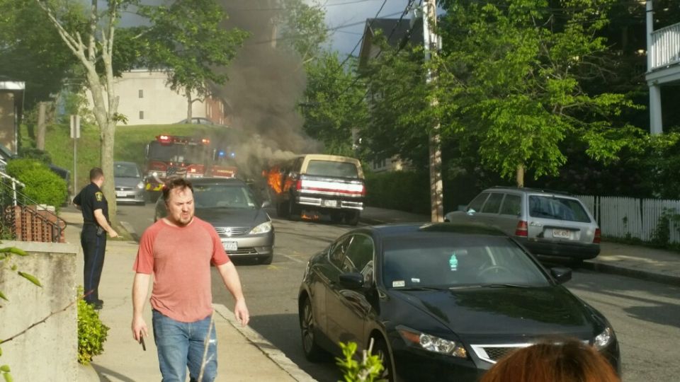 Van on fire with smoke billowing. Fire trucks and police are on scene. Several other cars are parked nearby