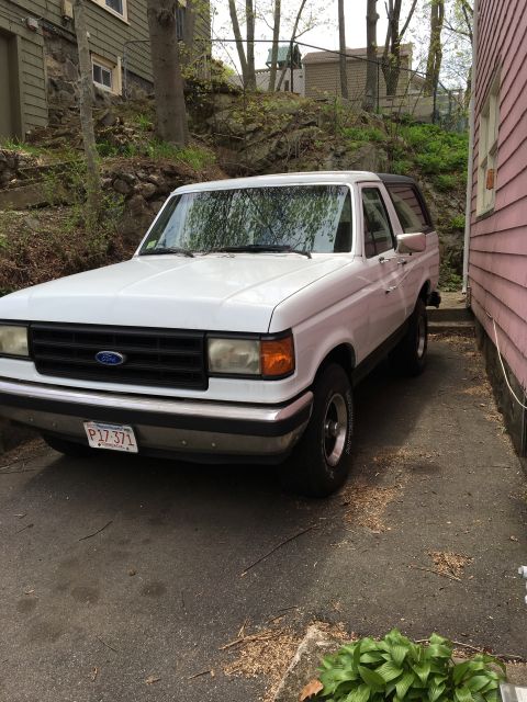 1990 Ford Bronco - A white Ford Bronco is parked in a driveway next to a pink house, with a wooded hill in the background.