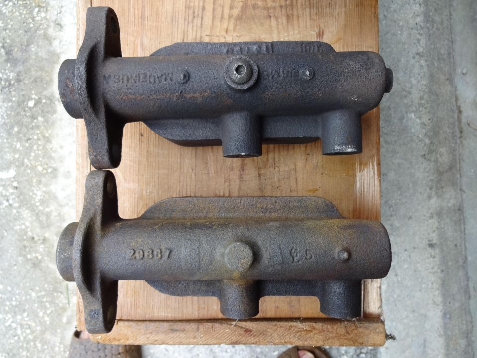 Two rusty cast-iron brake master cylinder castings on a wood board, one stamped 28867