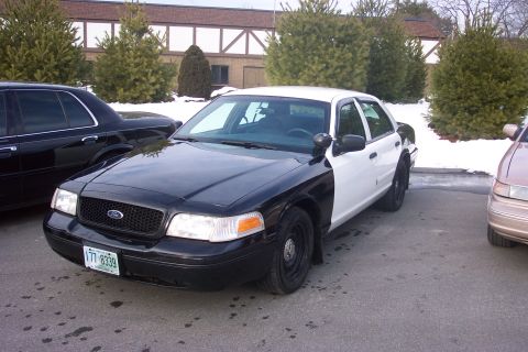 2001 Ford Crown Victoria - A two-tone police car is parked in a lot with other vehicles, with snow visible in the background.
