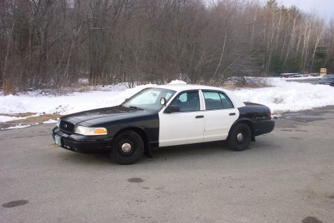 2001 Ford Crown Victoria - A two-tone police car, likely a 2001 Ford Crown Victoria, is parked in a lot with snow-covered trees in the background.