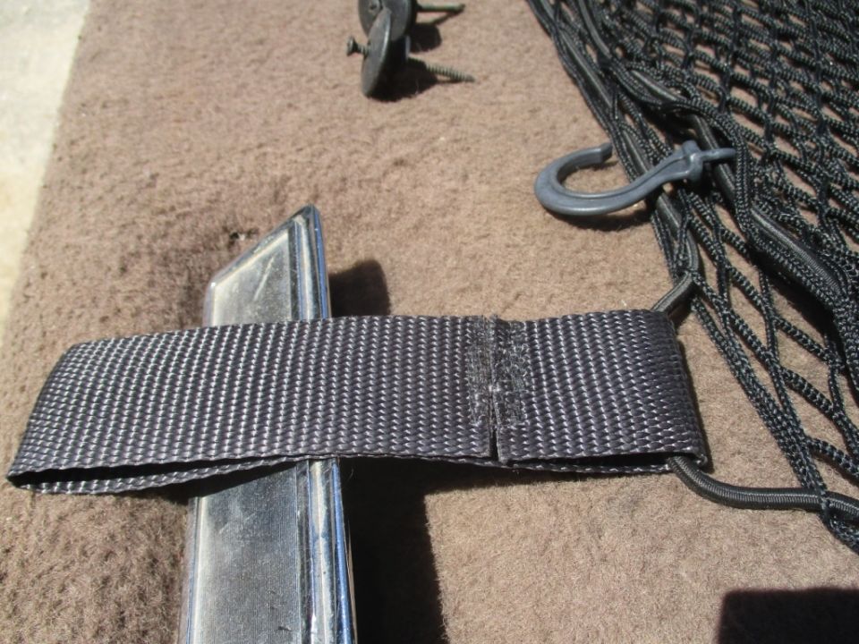 Cargo net with hook and strap attached to a metal trim piece on carpeted surface. Screws visible
