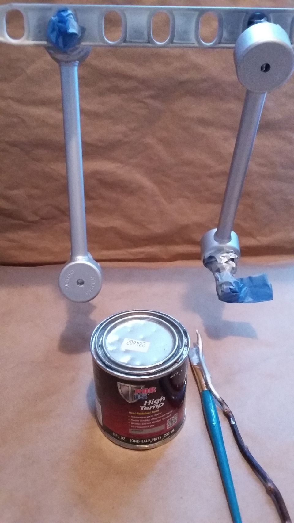 Two silver control arms hanging on a rack, with a can of High Temp paint and brushes below