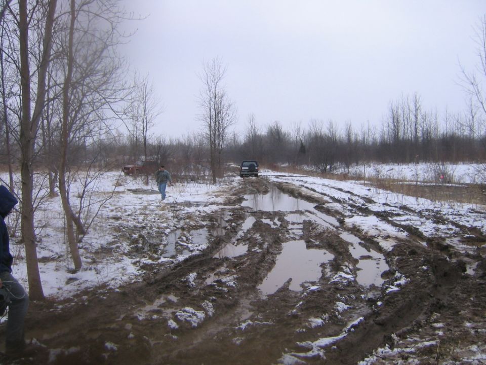 Trucks on a muddy rutted trail across a snowy field with bare winter trees
