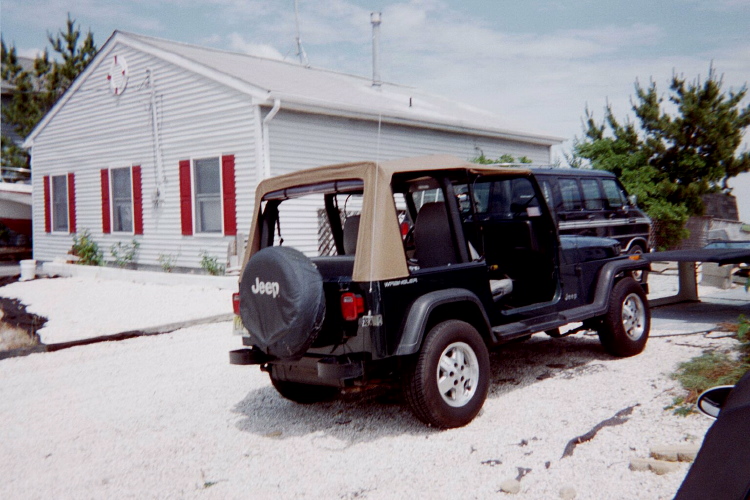 Black Jeep Wrangler with tan top parked next to a house. A black van is behind it
