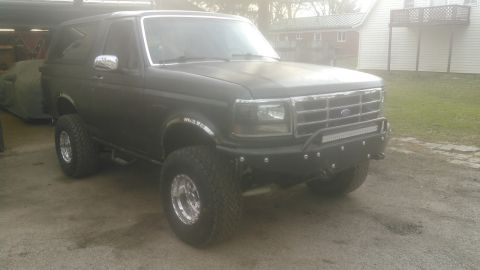 1992 Ford Bronco - A dark gray 1992 Ford Bronco is parked outside a building, modified with an aftermarket bumper and light bar.
