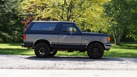 1987 Ford Bronco - A two-tone blue and gray 1987 Ford Bronco is shown from the side on a gravel surface with trees in the background.