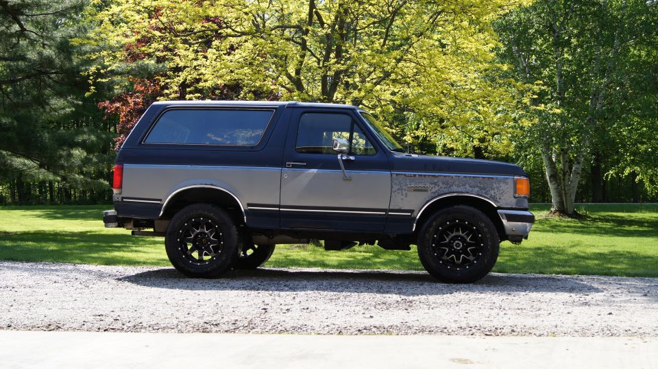 Two-tone blue and gray 1987 Ford Bronco with aftermarket black wheels parked on a gravel driveway