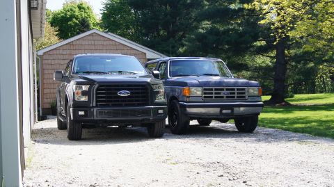 1987 Ford Bronco - A blue 1987 Ford Bronco and a black Ford F-150 are parked side-by-side in front of a garage and a tree.