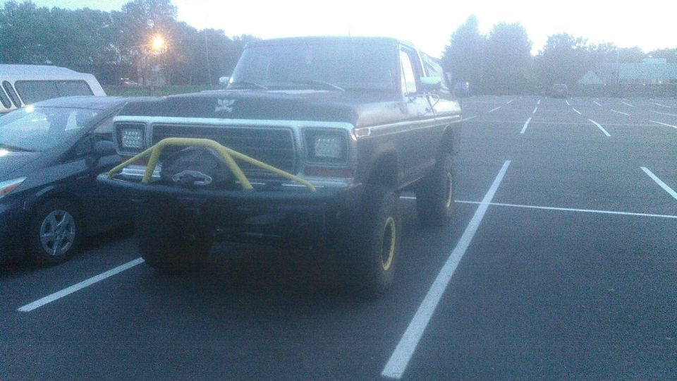 Lifted dark Ford Bronco with yellow brush guard and aftermarket headlights in a parking lot.