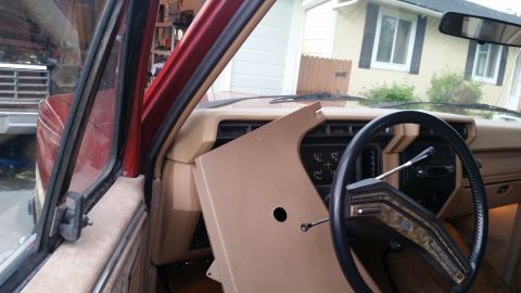 1985 Ford Bronco - Interior view of a red Ford Bronco showing the dashboard with a removed tan panel and the steering wheel. The exterior of the vehicle is vi…