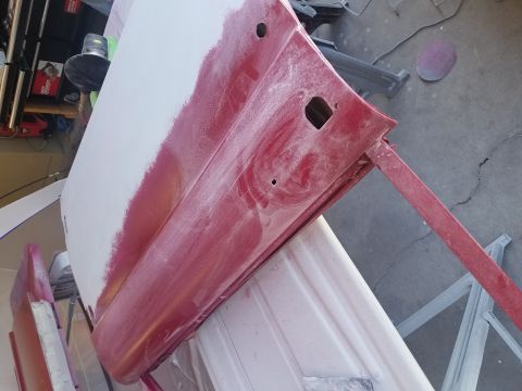 1968 Ford Bronco - A red car door is shown being prepped for paint, with primer and sanding dust visible. The door is mounted on a stand, with tools and other…