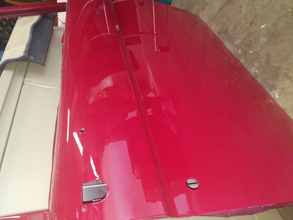 Shiny red vehicle door with silver latch mechanism laid on concrete next to cream body panel
