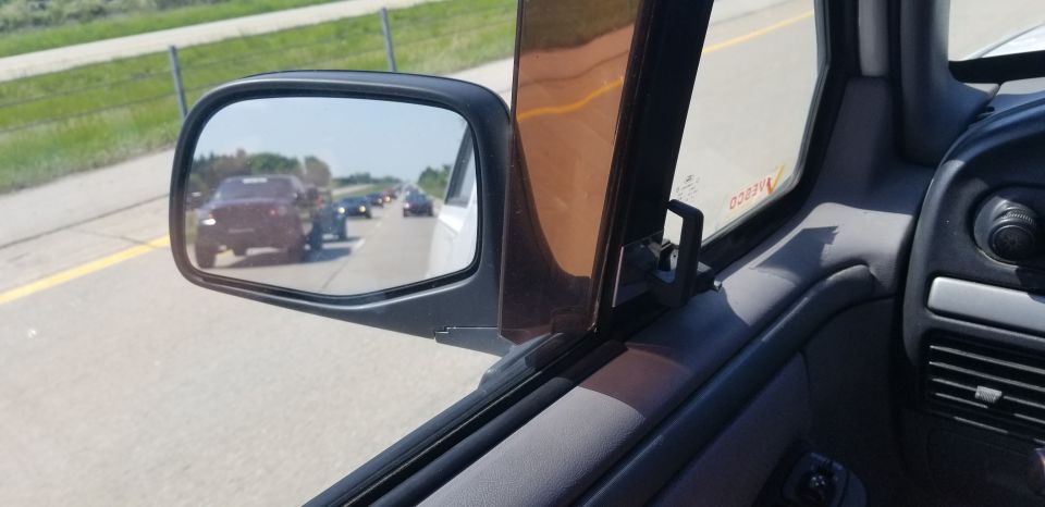 Driver's side mirror view of a dark SUV towing a trailer on a highway.