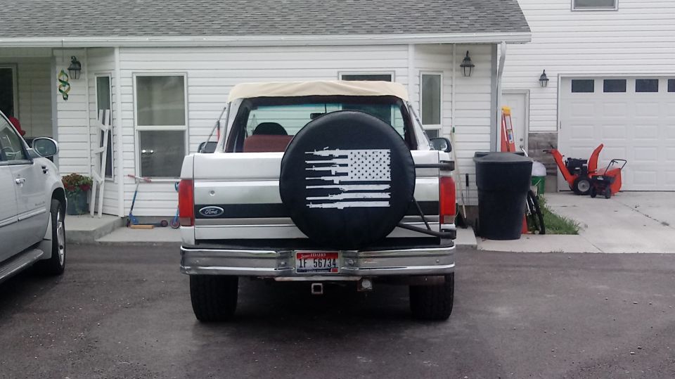 Rear view of a white 1995 Ford Bronco with a spare tire cover showing an American flag made of rifles.