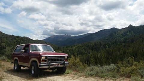 1985 Ford Bronco - A maroon and tan two-door Ford Bronco is parked on a dirt road with a mountainous, forested landscape in the background.
