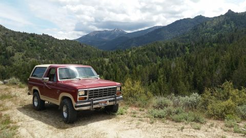 1985 Ford Bronco - A maroon and tan 1985 Ford Bronco is parked on a dirt road with a mountainous, forested landscape in the background.