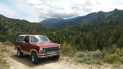 1985 Ford Bronco - A maroon and tan 1980s Ford Bronco is parked on a dirt road with a backdrop of pine-covered mountains under a cloudy sky.