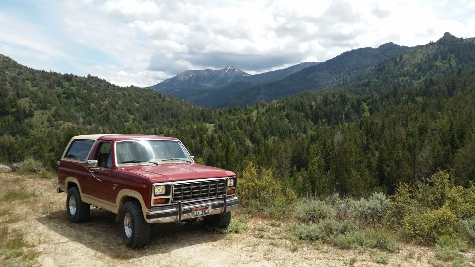 Maroon and tan 1980s Ford Bronco parked on dirt road, mountains in background.