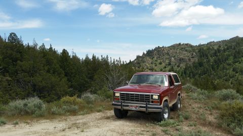 1985 Ford Bronco - A maroon 1985 Ford Bronco is parked on a dirt road in a natural landscape with trees and hills under a blue sky with clouds. The vehicle is…