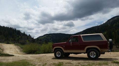 1985 Ford Bronco - A maroon and tan 1985 Ford Bronco is parked on a dirt road with a wooded hillside and cloudy sky in the background. The SUV is viewed from…