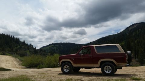 1985 Ford Bronco - A maroon and tan two-door SUV is parked on a dirt road with a wooded mountain range and cloudy sky in the background.
