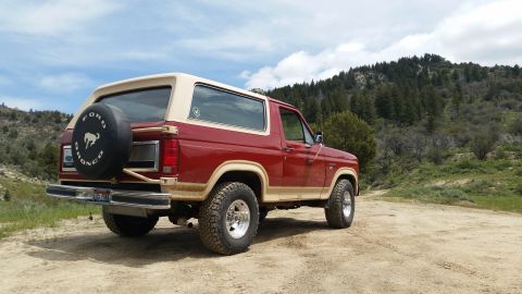 1985 Ford Bronco - A red and tan Ford Bronco is parked on a dirt road with a wooded hillside in the background. The rear of the vehicle is visible, including…