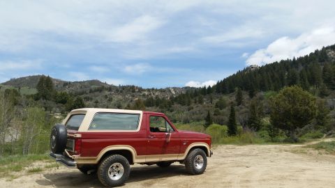 1985 Ford Bronco - A red and tan 1985 Ford Bronco is parked on a dirt road with a mountainous, tree-covered landscape in the background under a partly cloudy…