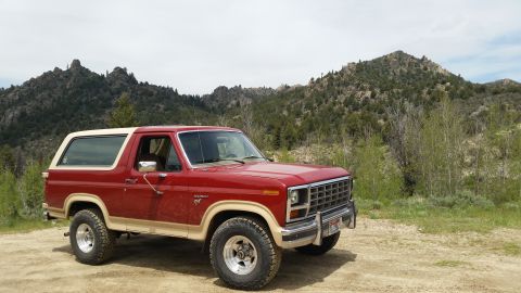 1985 Ford Bronco - A red and tan Ford Bronco is parked on a dirt road with a mountainous, wooded landscape in the background.