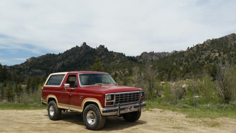 1985 Ford Bronco - A red and tan 1980s Ford Bronco is parked on a dirt road with a mountainous, tree-covered landscape in the background.