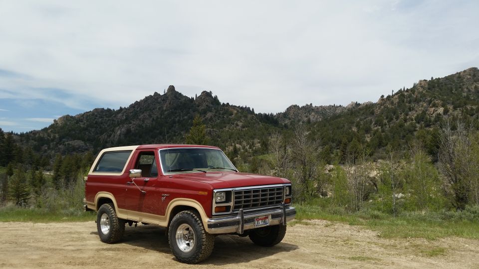Red and tan 1980s Ford Bronco parked on dirt road with mountains in background.