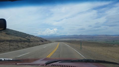 1985 Ford Bronco - View from the driver's seat of a maroon vehicle driving on a highway through a dry, hilly landscape under a cloudy sky. The road ahead has…