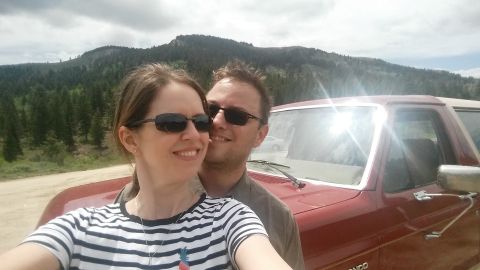 1985 Ford Bronco - A couple takes a selfie in front of a red Ford Bronco with a mountainous, tree-covered landscape in the background.