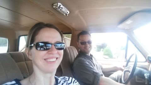 1985 Ford Bronco - A man and a woman smile at the camera from inside a vehicle, likely a 1985 Ford Bronco, as they appear to be on a road trip. The woman is i…