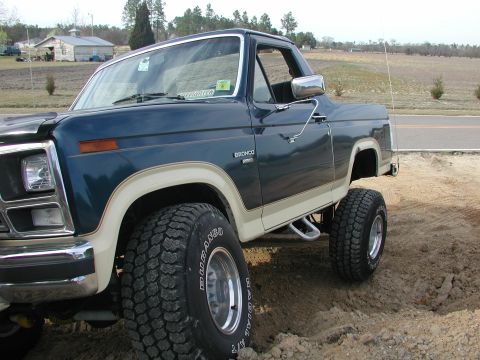 1985 Ford Bronco - This lifted Ford Bronco, likely from the early 1980s, sits on a dirt lot with a rural background.