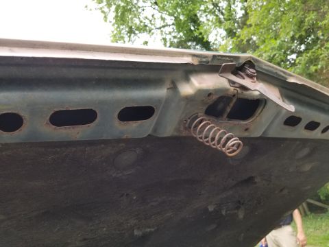 2001 Ford Crown Victoria - The underside of a car hood shows a rusty latch mechanism and spring, with oval cutouts in the metal.