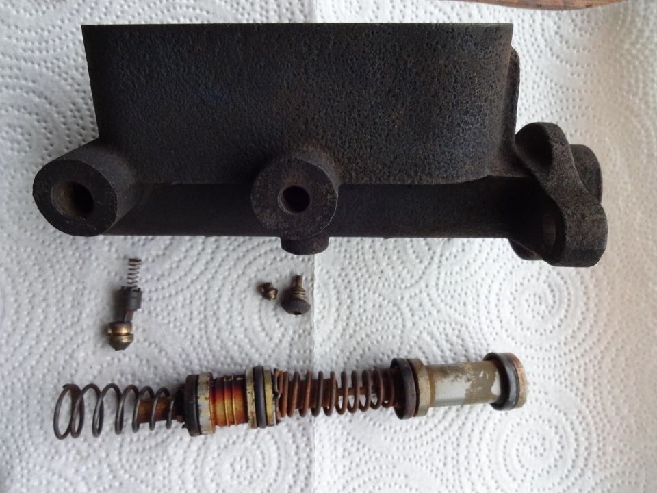 Disassembled brake master cylinder and internal parts laid out on paper towels. Includes cylinder body, piston, spring, and small hardware
