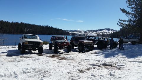 1968 Ford Bronco - A group of off-road vehicles are parked in the snow near a lake on a clear day.