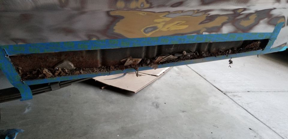 Cut-open rusty rocker panel with debris inside, taped with blue masking tape on a primered car in a garage