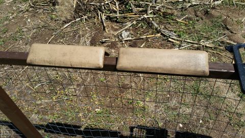 1985 Ford Bronco - Tan armrests are mounted on a metal bar above a wire fence, with dry vegetation in the background.