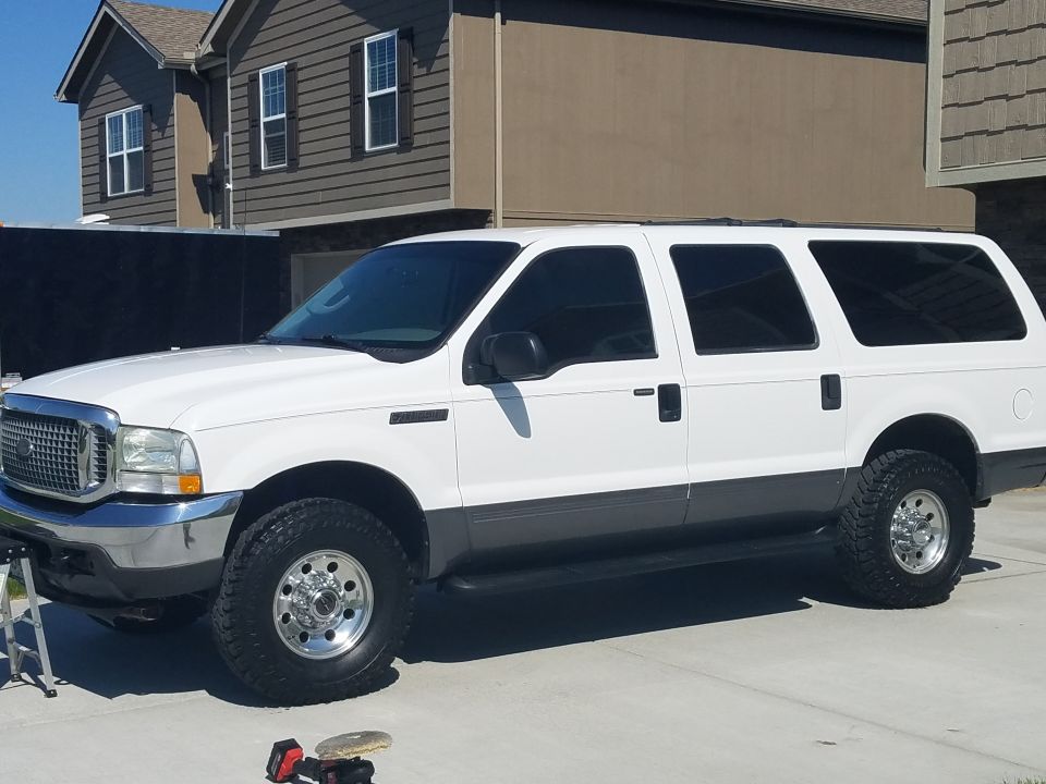 White Ford Excursion SUV, driver-side front angle, on driveway with mud tires and polished wheels