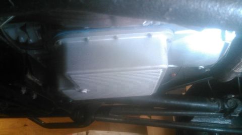 1992 Ford Bronco - The underside of a vehicle shows a silver transmission pan with fins and a drain plug. The pan is attached to the transmission with several…
