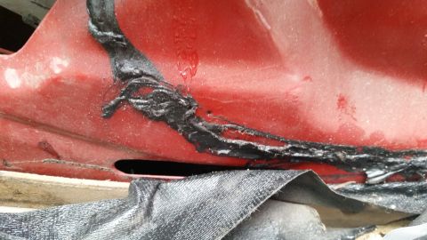 1985 Ford Bronco - A close-up view shows a section of a red vehicle body with a black sealant applied over a crack or seam. A piece of black mesh fabric is vi…